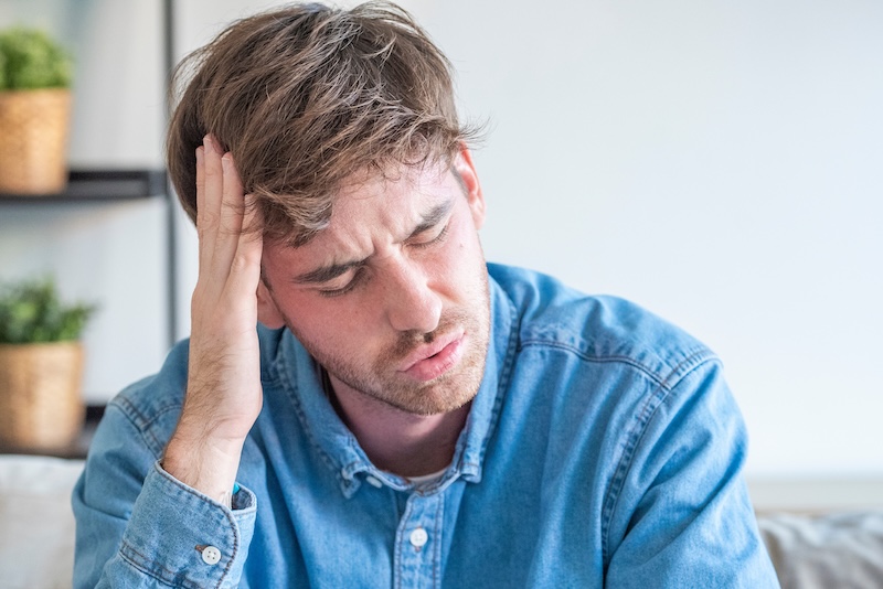 Young man holding his head in pain sitting on a couch, stressed and suffering from a headache or migraine at home, feeling tired and anxious
